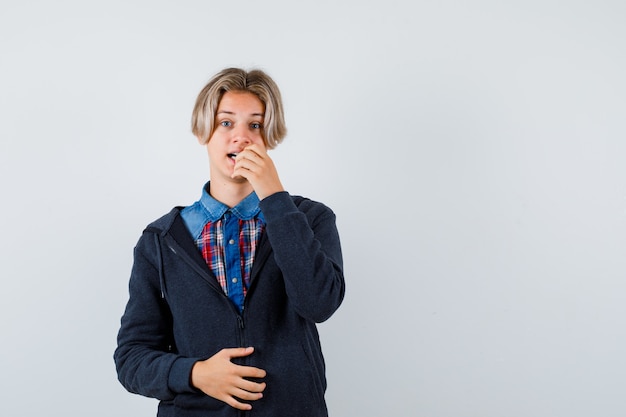 Cute teen boy in shirt, hoodie keeping hand on mouth and looking forgetful , front view.