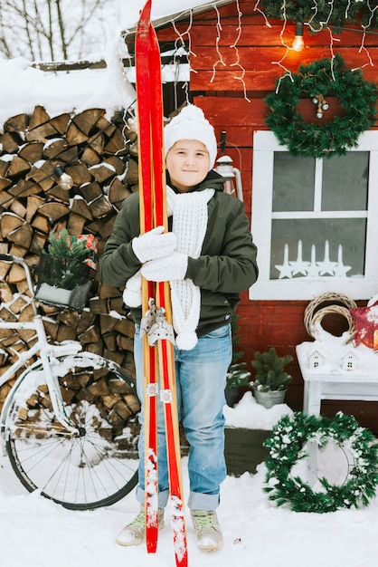 cute teenage boy in knitted white hat stand at porch of country house and decorate Christmas tree fo