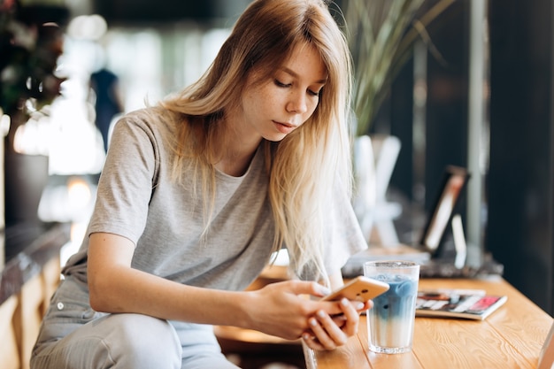 A cute thin blonde girl,dressed in casual style,drinks coffee and looks at her phone in a coffee shop. .