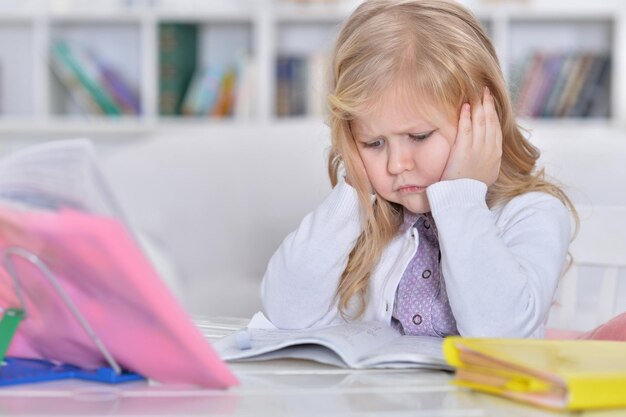 Cute tired schoolgirl sitting at table and doing homework at home