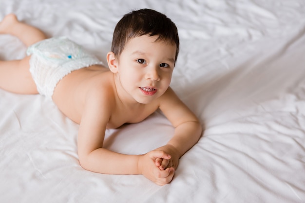 Cute toddler boy in diapers lies on bed with white sheet