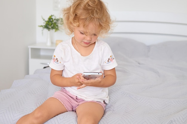 Photo cute toddler girl holding smartphone sitting on bed using digital device while playing online game browsing internet on cellphone scrolling on mobile phone in cozy bedroom