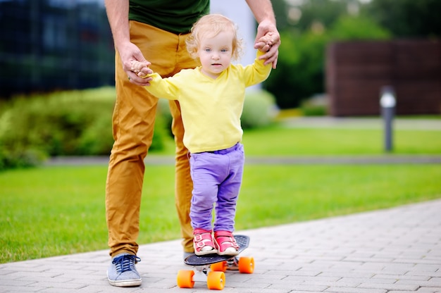 Cute toddler girl learning to skateboard with her father outdoors