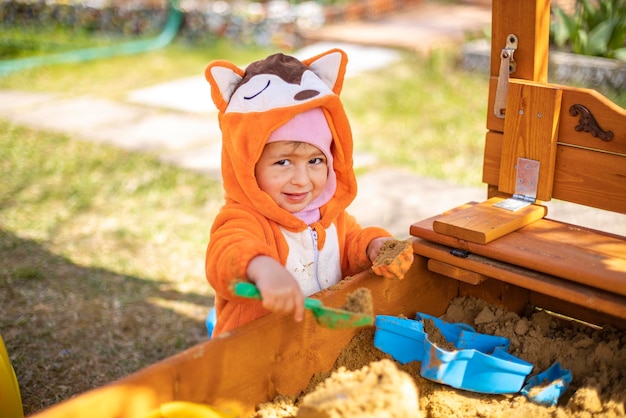 Cute toddler in orange jumpsuit plays in the sand in the sandbox outdoors on a sunny day