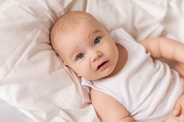 Cute toddler in white bodysuit lies in bed with white bedding