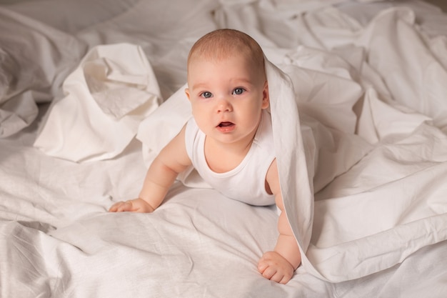 Cute toddler in white bodysuit lies in bed with white bedding