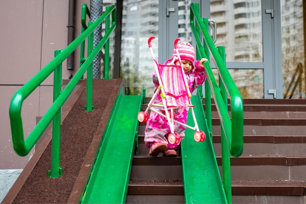 Cute toddler with a toy stroller walks along steel railing ramp for wheelchair carts and strollers gentle descent from the stairs on the street