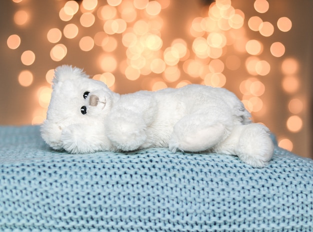 Cute white little teddy bear lying peacefully on light blue knitted blanket. Cozy sweater 