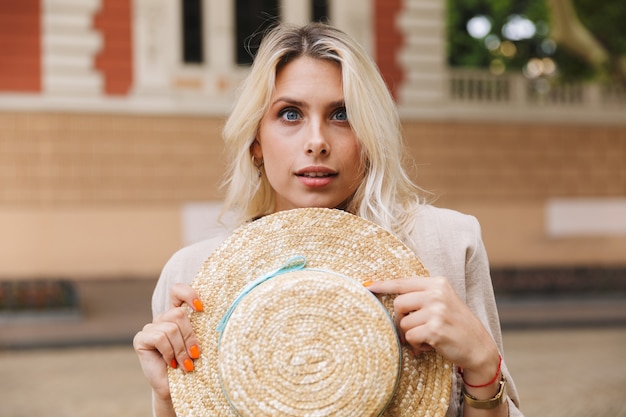 cute woman 20s holding straw hat, while walking through city street