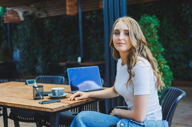 A cute woman drinks coffee on the terrace of a summer cafe and works on a laptop remote work while on vacation in a cafe with a laptop