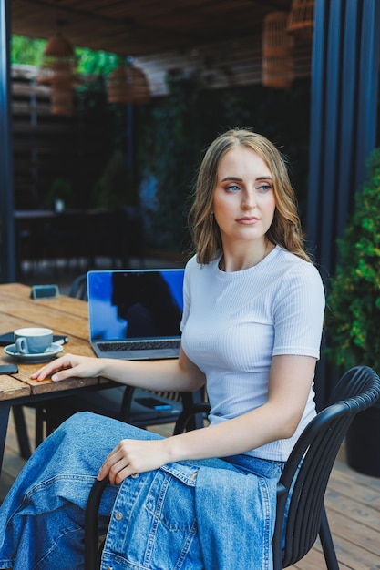 A cute woman drinks coffee on the terrace of a summer cafe and works on a laptop remote work while on vacation in a cafe with a laptop