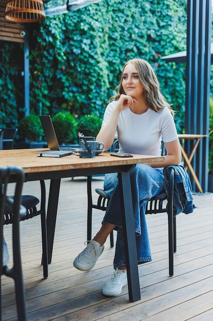 A cute woman drinks coffee on the terrace of a summer cafe and works on a laptop remote work while on vacation in a cafe with a laptop