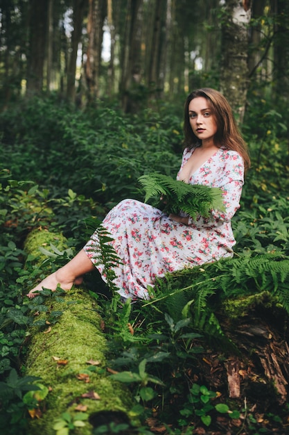 A cute woman in a floral dress is sitting with a fern bouquet in the forest.