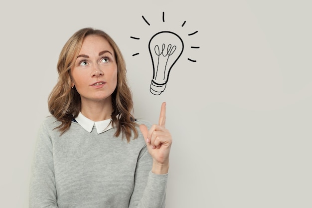 Cute woman in gray sweater pointing on light bulb idea on grey background