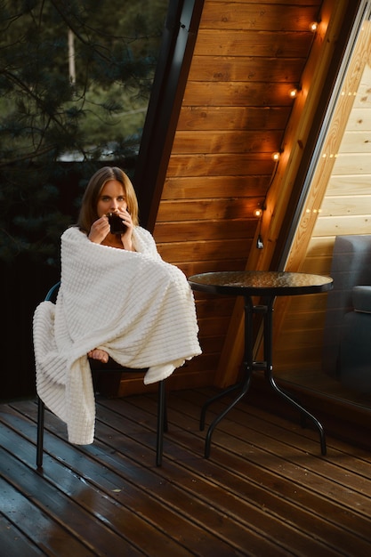 Cute woman is sitting in an armchair on the terrace of a country house wrapped in a blanket with a cup of soothing tea and enjoying the fresh air