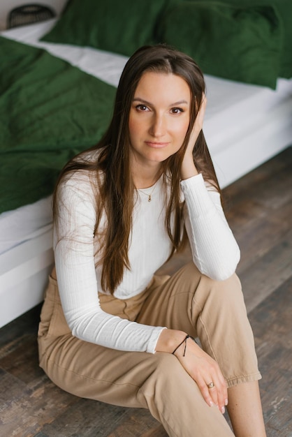 Cute woman is sitting on the floor near the bed and smiling at the camera at home