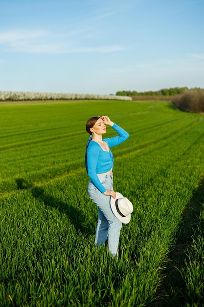 A cute woman in jeans and a hat stands in a green field A smiling woman in a blue top and jeans walks in the green grass