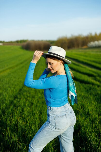 A cute woman in jeans and a hat stands in a green field A smiling woman in a blue top and jeans walks in the green grass