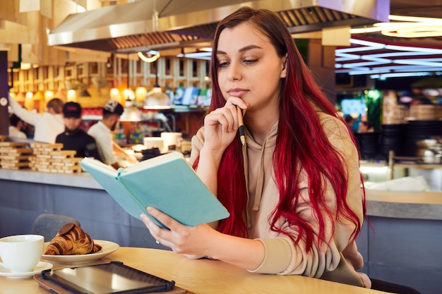 Cute woman with reddyed hair works remotely in a cafe holding a notebook in her hands