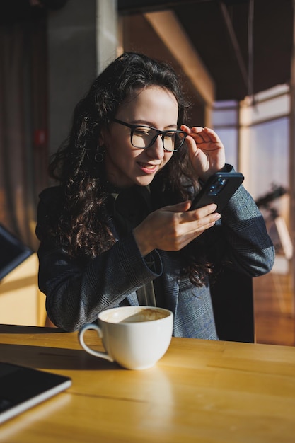 A cute woman works on a laptop in a cafe Young concentrated brunette woman in glasses sitting at the table near the window drinking coffee Freelance and remote work Modern women's lifestyle