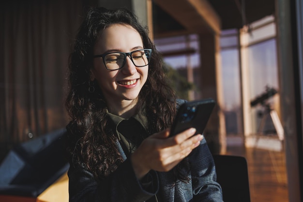 A cute woman works on a laptop in a cafe Young concentrated brunette woman in glasses sitting at the table near the window drinking coffee Freelance and remote work Modern women's lifestyle