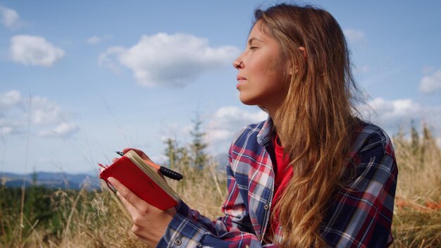 Cute woman writing notes in notebook with pencil Relaxed girl painting sketch of mountains at vacation Thoughtful female hipster looking away in field Young lady taking break outdoor