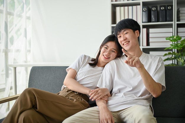 A cute young Asian girlfriend is hugging her boyfriend while enjoying watching TV on a couch