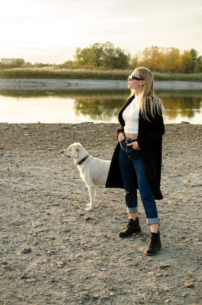 Cute young beautiful woman standing posing with her white retrieve dog on the river bank fulllength