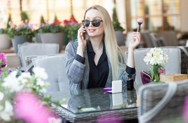 Cute young blonde woman holding a makeup brush in her hand and talking on phone while sitting in summer terrace of cafe