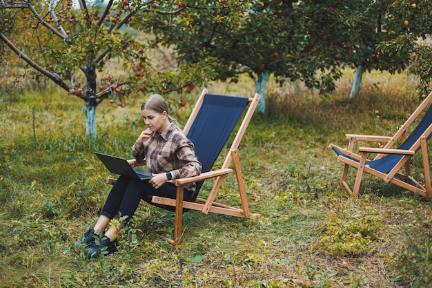 A cute young female freelancer is working in the garden outdoors while sitting in a comfortable garden chair Remote work on a portable laptop