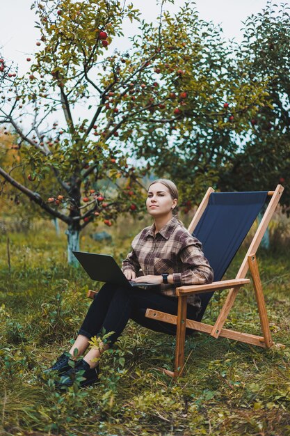 A cute young female freelancer is working in the garden outdoors while sitting in a comfortable garden chair Remote work on a portable laptop