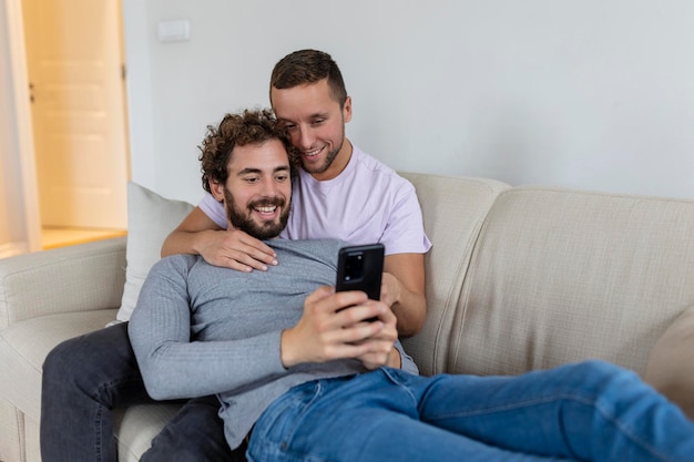 Cute young gay couple video calling their friends in their living room at home Two male lovers smiling cheerfully while greeting their friends on a smartphone Young gay couple sitting together