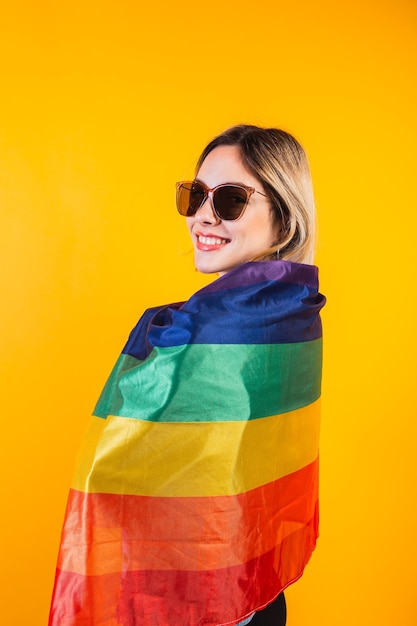 Cute young girl drapes herself with large rainbow flag on yellow background.