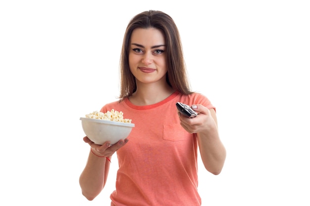 Cute young girl looks forward and holds the remote and the plate with the popcorn is isolated on a white background