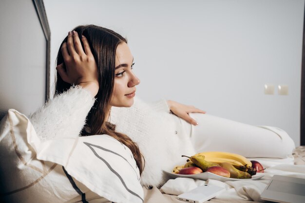 Cute young girl in pajamas lies in bed in the evening watching an interesting TV show on her laptop