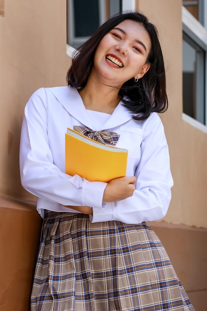 Cute and young girl wearing Japanese, Korean style schoolgirl uniform holding book and pose to camera with fun and happy in front of school building.