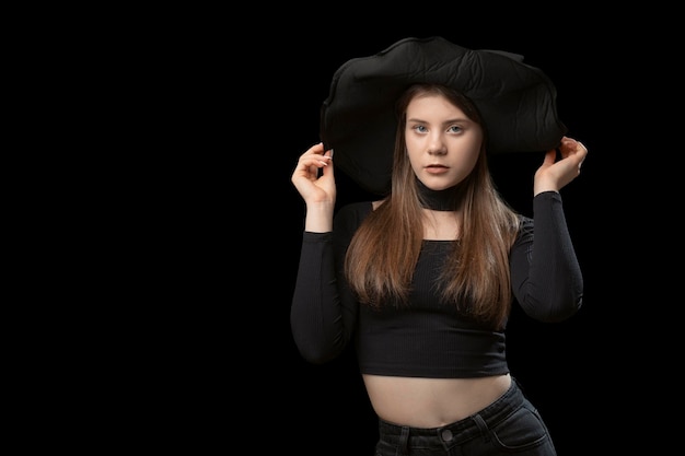 Cute young girl wears tightfitting black top and widebrimmed hat Studio portrait on black background Copy space