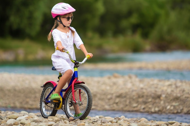 Cute young girl in white clothing, sunglasses with long braids wearing pink safety helmet riding child bicycle on pebbled river bank on blurred green summer. Outdoors activity concept.