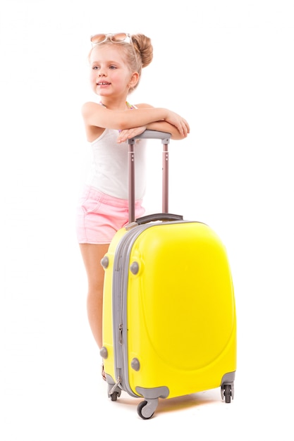 Cute young girl in white shirt, pink shorts and sunglasses stand near yellow suitcase