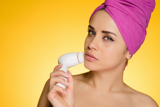 Cute young girl with a pink towel on her head doing a deep cleansing of the skin of the face with an electric brush