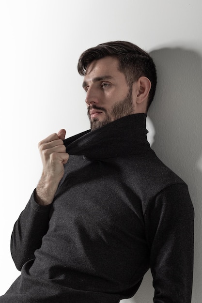 Cute young guy standing by the white wall and touching the collar of his poloneck