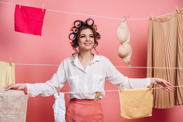 Cute, young housewife on a wall of ropes with clothes on a pink background