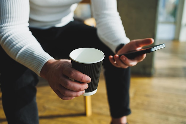 Cute young male freelancer drinking coffee while sitting in bright workspace working remotely A man in a white sweater and trousers works happily in the office