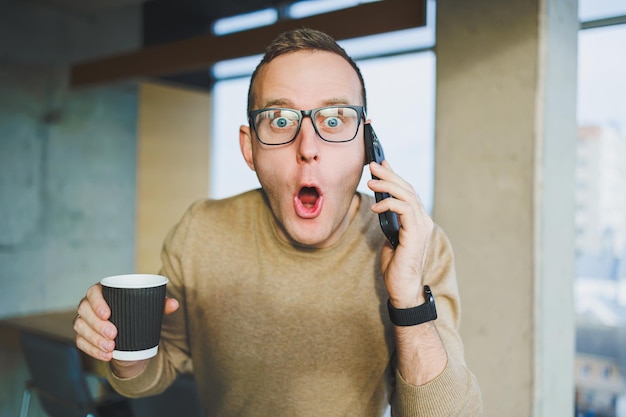 A cute young man in a brown sweater and glasses is talking emotionally on a mobile phone and drinking coffee while relaxing in the office A young freelancer works remotely