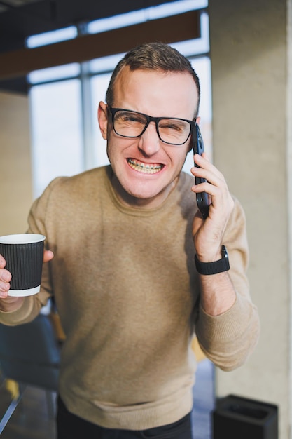 A cute young man in a brown sweater and glasses is talking emotionally on a mobile phone and drinking coffee while relaxing in the office A young freelancer works remotely