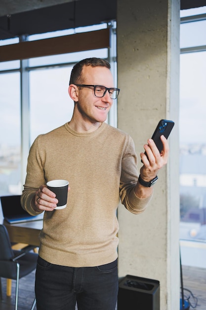 A cute young man in a brown sweater and glasses is talking emotionally on a mobile phone and drinking coffee while relaxing in the office A young freelancer works remotely