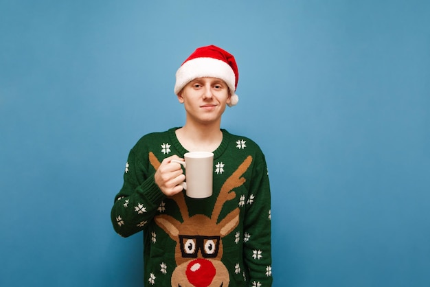 Cute young man in christmas clothes standing with cup of hot drink on blue background