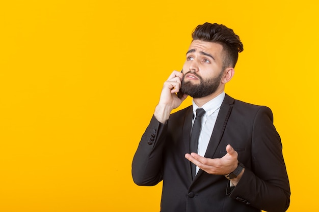 Cute young man with a beard in formal clothes talking on the phone posing on a yellow wall