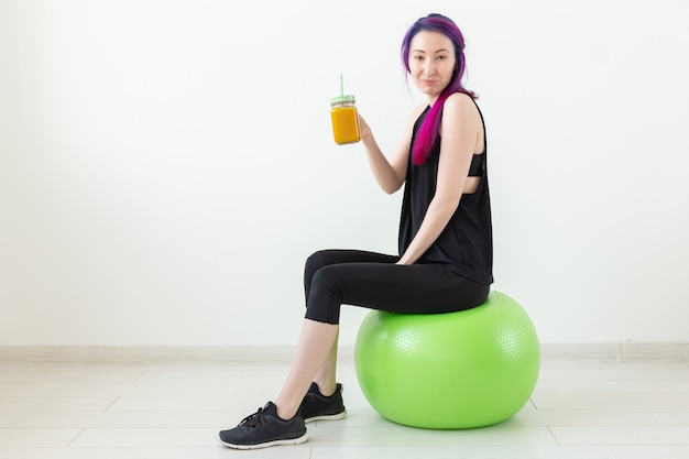 Cute young mixed race hipster girl with colored hair sitting on a green fitball and holding a banana protein smoothie in her hands on a white background. Healthy eating and exercise concept. Copyspace