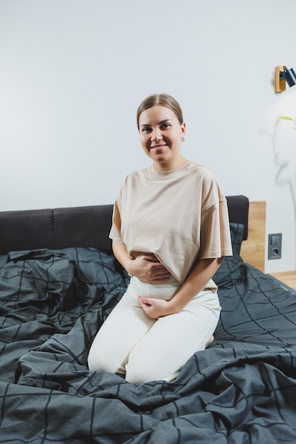 Cute young pregnant woman sitting on bed with gray sheets in casual clothes Cute pregnant woman at home